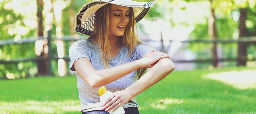 Mulher jovem loira com chapéu passa protetor solar enquanto está sentada em parque